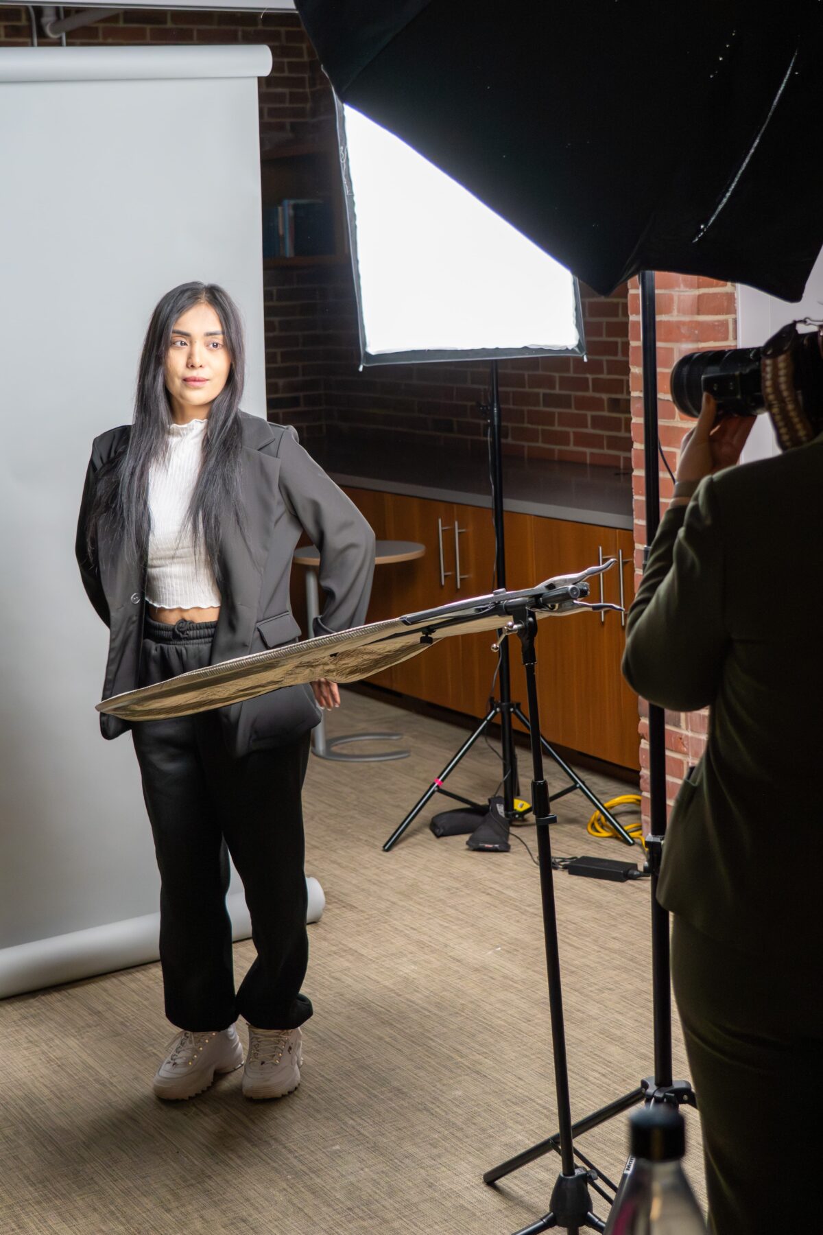 Mia Isabella – Adelphi BTS Behind the scenes photography by Lens with Benefits captured at Adelphi University. A young woman in a gray oversized blazer, white crop top, and black trousers poses against a white backdrop while a photographer frames the shot. Professional studio lighting equipment including two softboxes and a gold reflector panel are visible in the foreground. Exposed brick walls and wooden cabinetry in the background. Editorial behind the scenes photography for NYC-based photographer Mia Isabella.