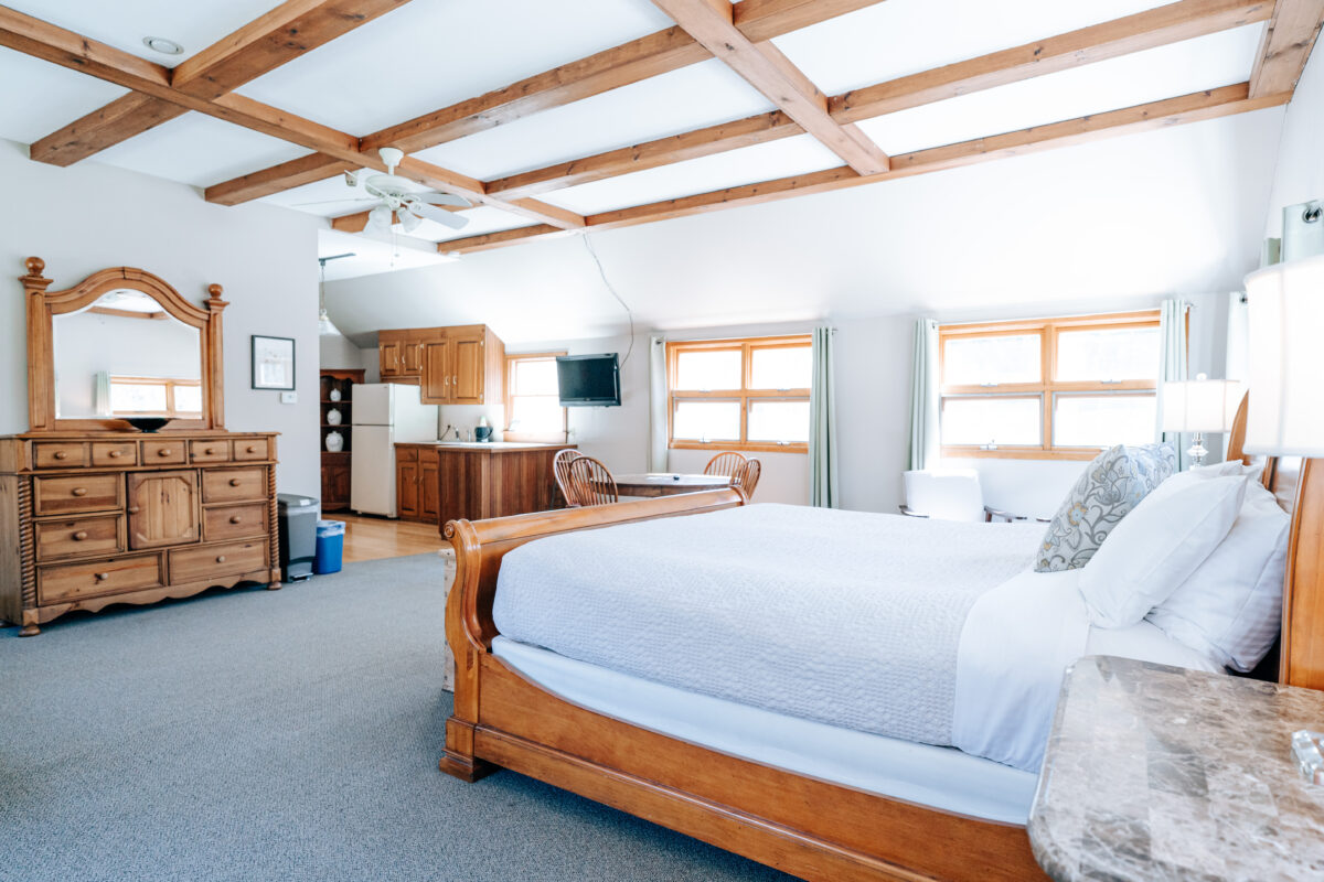 Hollyhedge Estate room Commercial interior photography by Lens with Benefits of a guest suite at HollyHedge Estate in New Hope, Pennsylvania. A spacious open-plan room features a carved wood sleigh bed with white bedding, exposed wood coffered ceiling beams, a kitchenette with wooden cabinetry and full-size refrigerator, a large pine dresser with mirror, and multiple windows flooding the space with natural light. Boutique inn and hospitality photography in Bucks County.