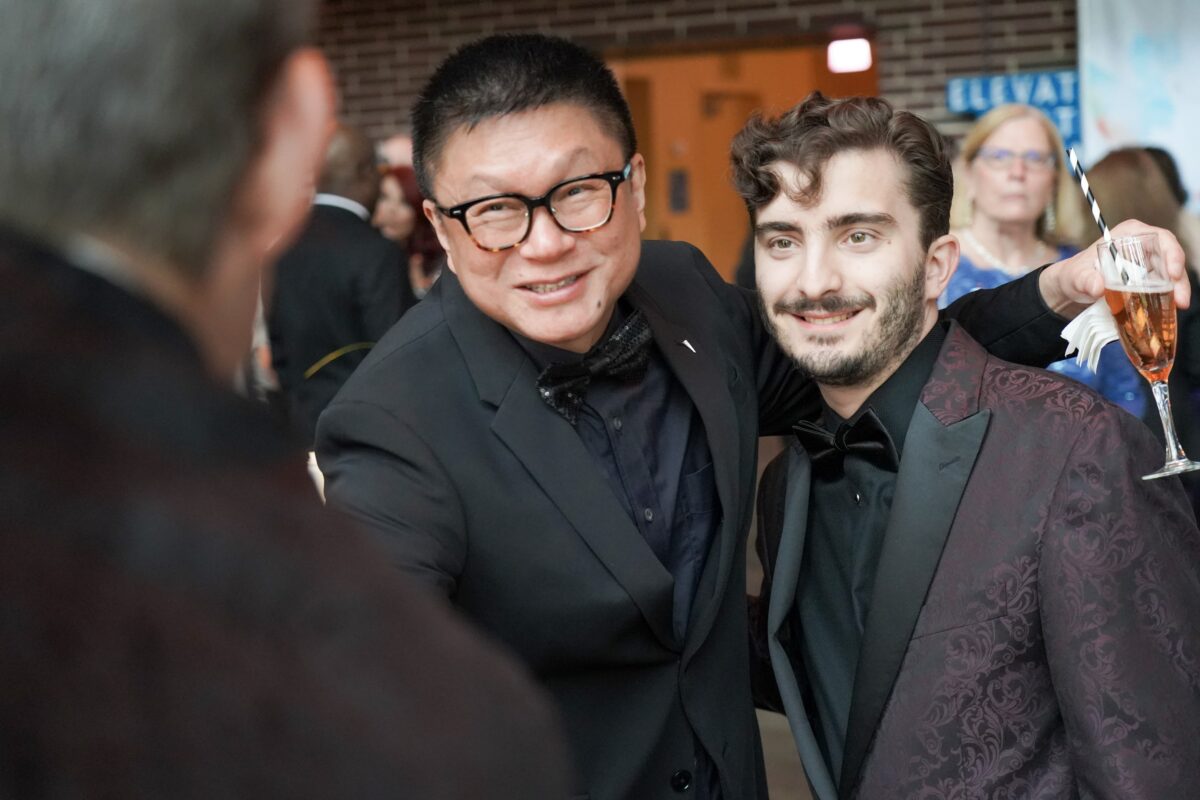 Two River Theater Company gala Candid event photography captured by Lens with Benefits at the Two River Theater Company's annual gala and fundraiser in New Jersey. Two formally dressed attendees in black tuxedos share a warm, laughing moment amid a crowded reception hall. The subject on the right holds a glass of rosé. Brick wall and event signage visible in the background. Editorial event and nonprofit photography.