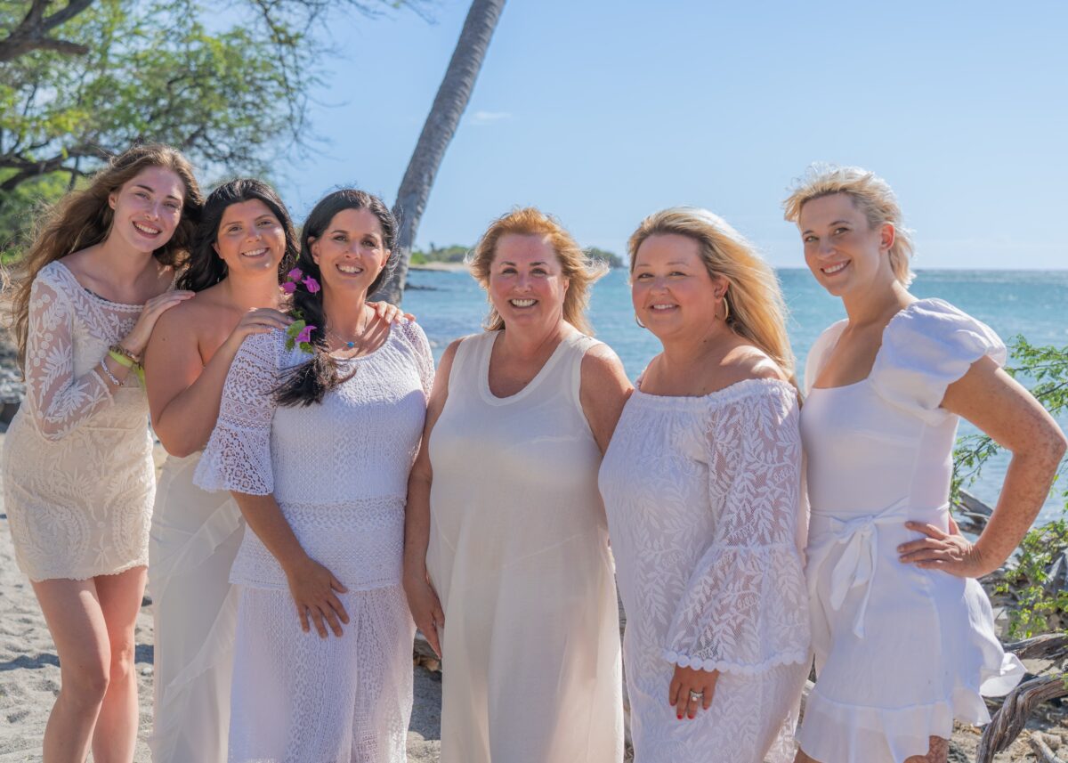 A family in white on a beach in Hawaii. Outdoor family portrait photography by Lens with Benefits at Waikoloa Beach on the Big Island of Hawaii. Six women dressed in white and cream lace stand together on a sandy beach with turquoise ocean water, a leaning palm tree, and clear blue sky in the background. Candid lifestyle family photography capturing a multi-generational group celebrating a 70th birthday milestone.